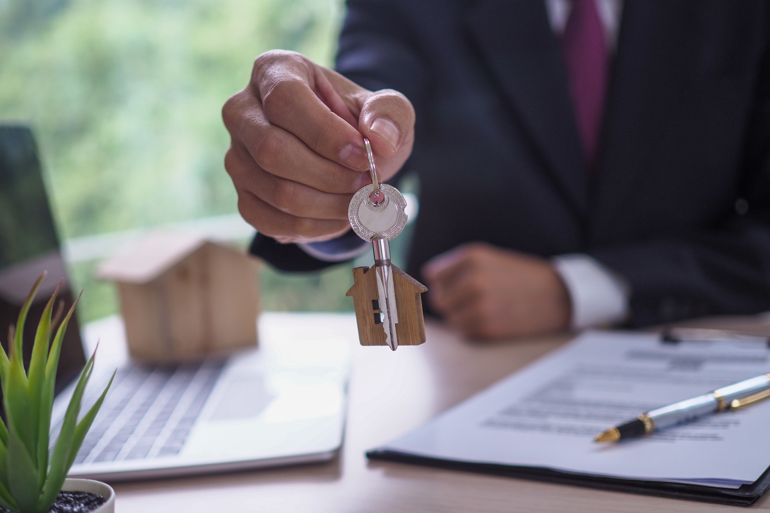 A hand passing a key over a desk featuring a laptop and scattered papers, symbolizing a property handover or real estate transaction.