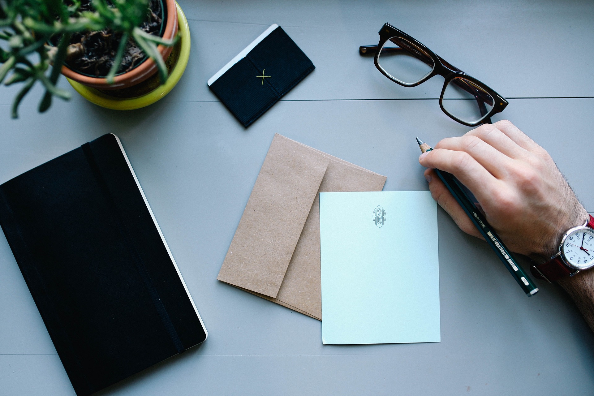 A flat-lay of a workspace featuring a hand holding a pencil, a black notebook, brown envelopes with a lien letter, a light blue card, glasses, a plant in a yellow pot, and a black cardholder. The setting conveys a clean and organized desk for writing or planning.