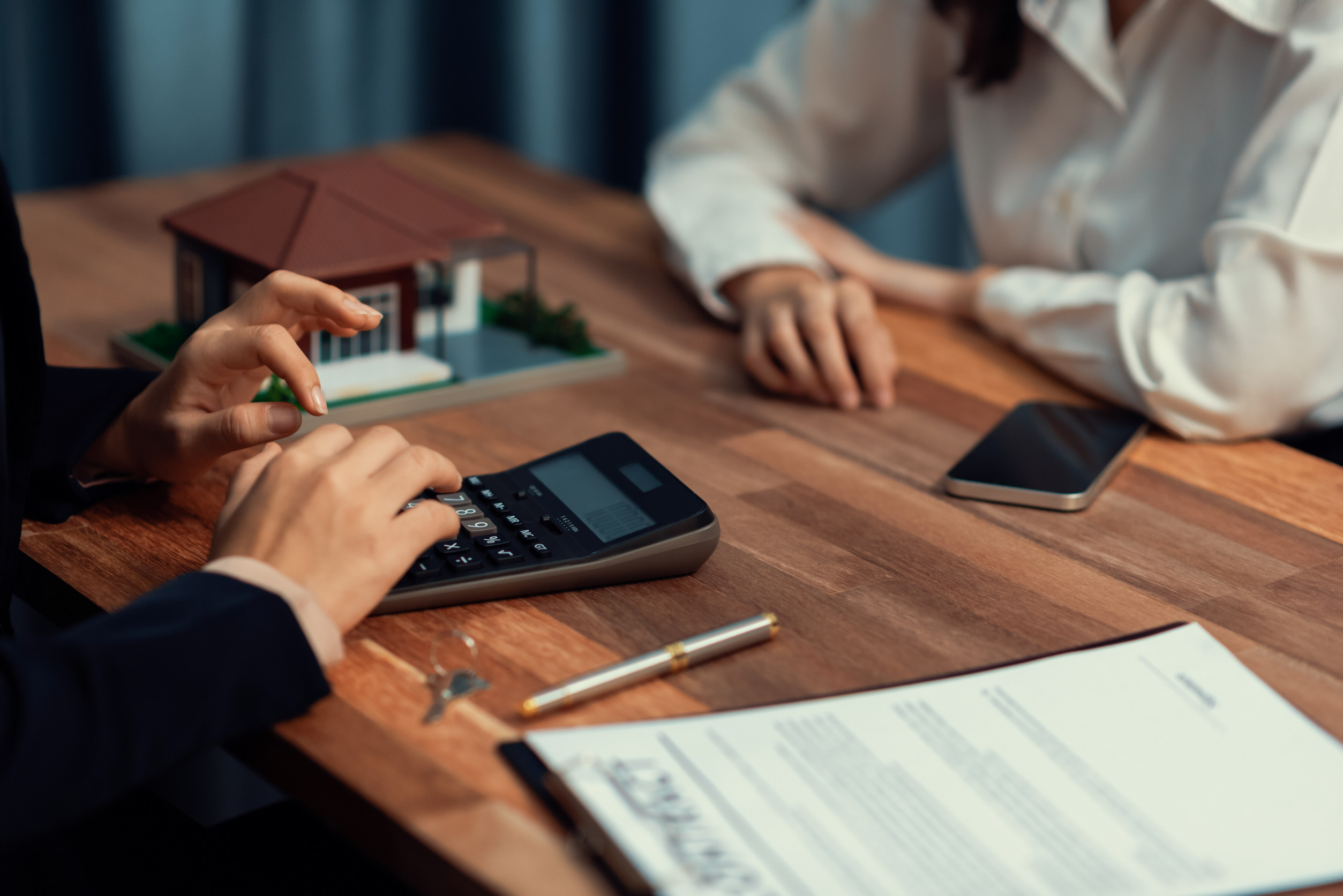 A close-up of a real estate transaction, showing a person using a calculator while another individual sits across the table. A model house, contract, pen, keys, and a smartphone are visible on the wooden desk, indicating a discussion related to property purchase or financial calculations doing a special assessments.